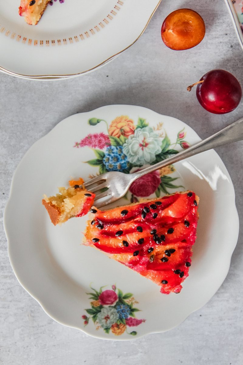 A slice of upside down plum passionfruit cake sits on an individual plate with a forkful of cake sitting beside on a gray surface.