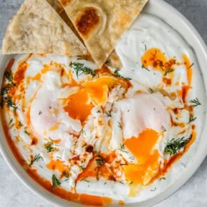 Turkish Cilbir - Poached Eggs in Yogurt sit broken in a rimmed ceramic plate with a few pieces of bread served on the side on a gray surface.