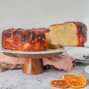 A slice of Greek Orange cake sits on a serving knife above a cake stand on a gray surface.