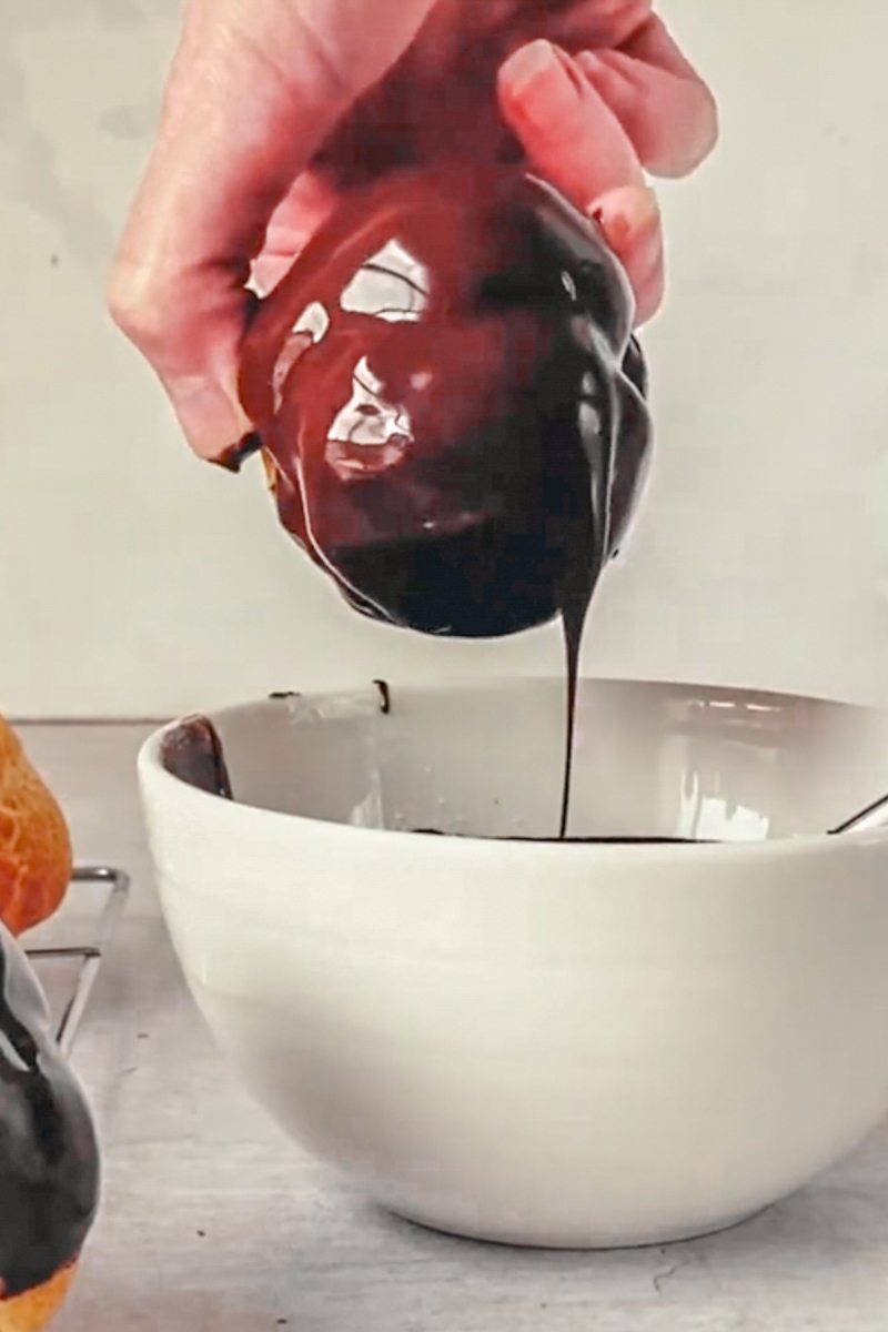 A choux pastry dipped into melted chocolate has chocolate dripped off from a white ceramic bowl on a gray surface.