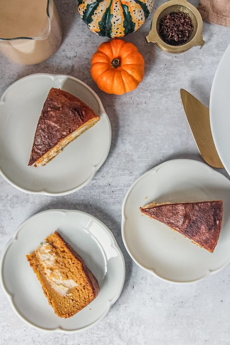 Slices of pumpkin cake with a cheesecake center are served up on white cake plates on a gray surface.