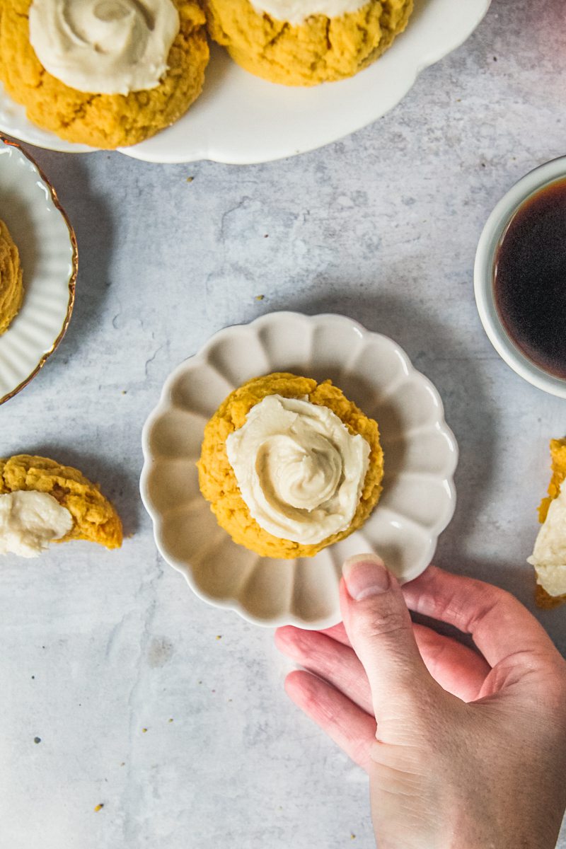 A pumpkin sugar cookie sits on an individual fluted gray ceramic plate that is being picked up with maple buttercream frosting in the center above a gray surface.