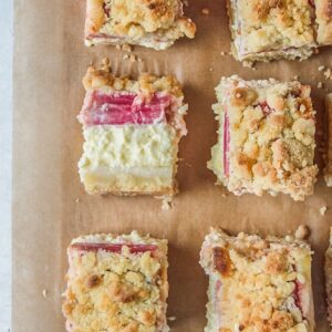 A square of Estonian rhubarb crumble cheesecake lays on its side showing the layers of cake whilst a separate square is picked up from parchment paper.