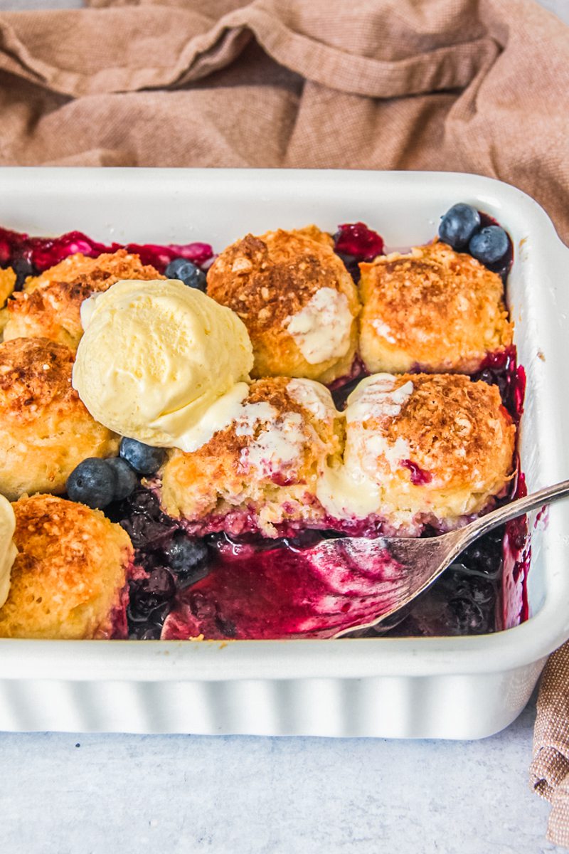 A blueberry cobbler sits in a white ceramic baking dish on a gray surface with a brown cloth behind it.