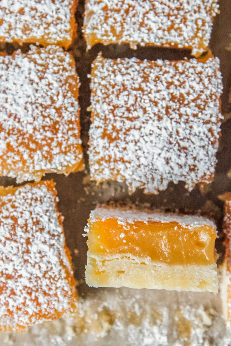 Lemon shortbread bars sit on a parchment paper lined surface with one sitting on their side showing the shortbread base and lemon curd topping visible.