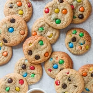 M&M cookies lay unevenly on a gray surface with a cup of tea beside.