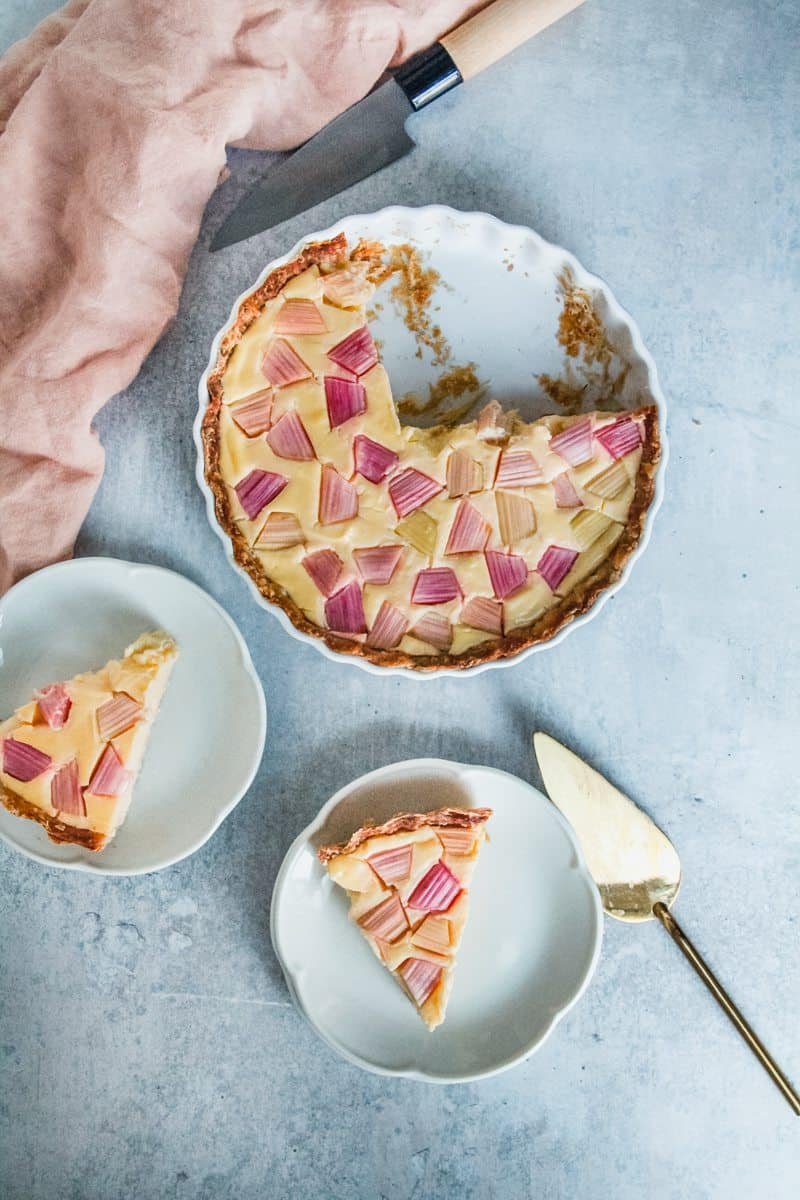 A rhubarb custard tart sits on a grey background with two slices served up on plates.