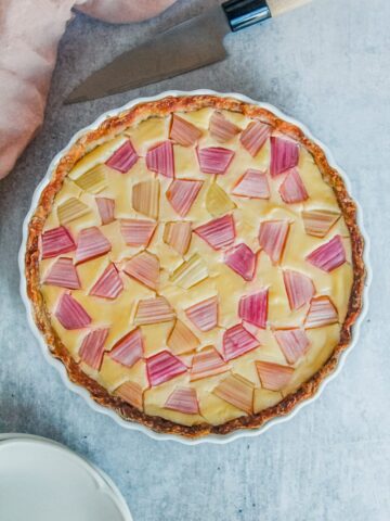A rhubarb custard tart with rough geometric rhubarb sits in a white ceramic baking dish on a gray surface.