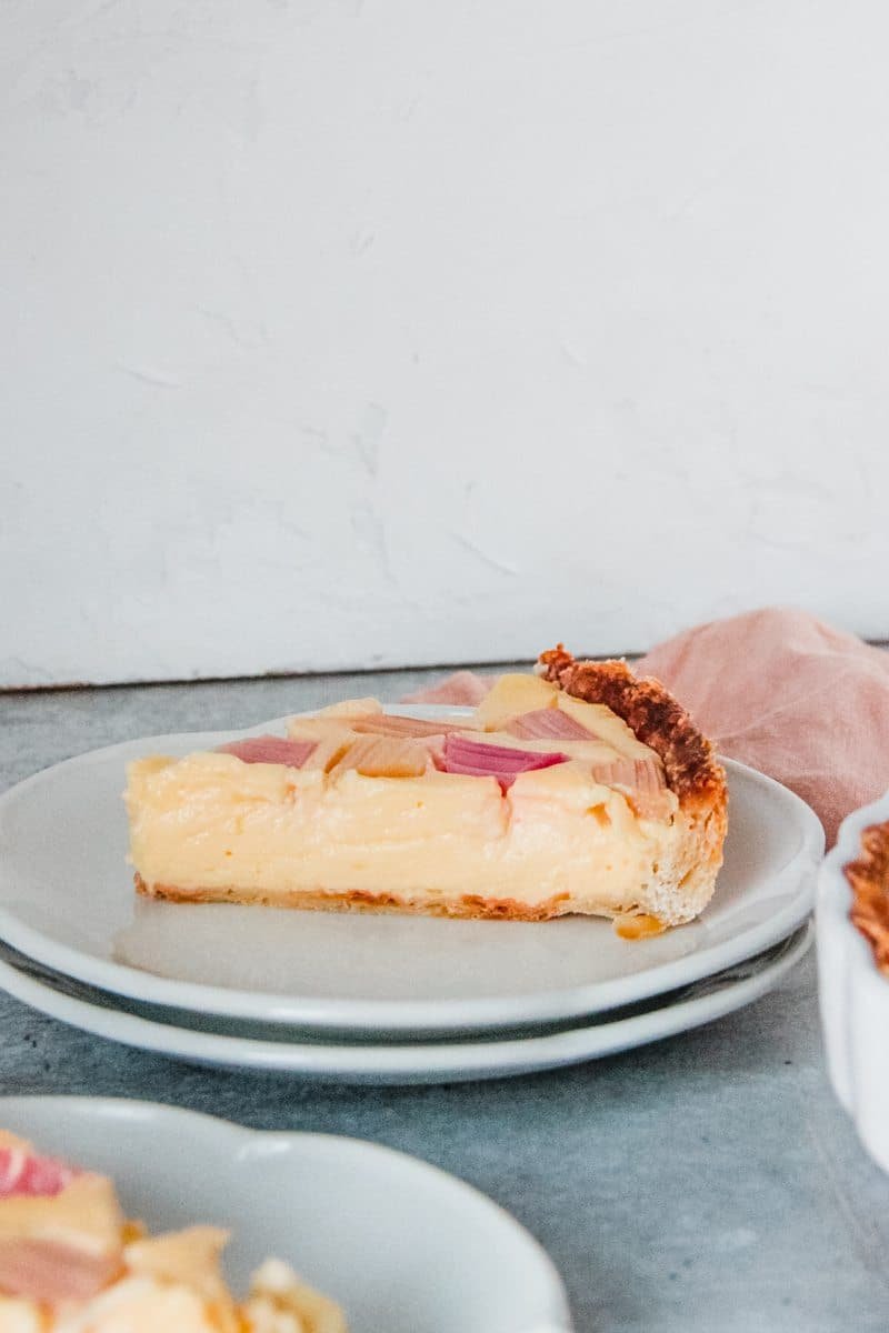 A piece of lemon custard tart sits served up on two plates with the flaky pastry profile visible and the lemon custard and pieces of rhubarb that are poking out of the top of the custard, sitting on a grey background.