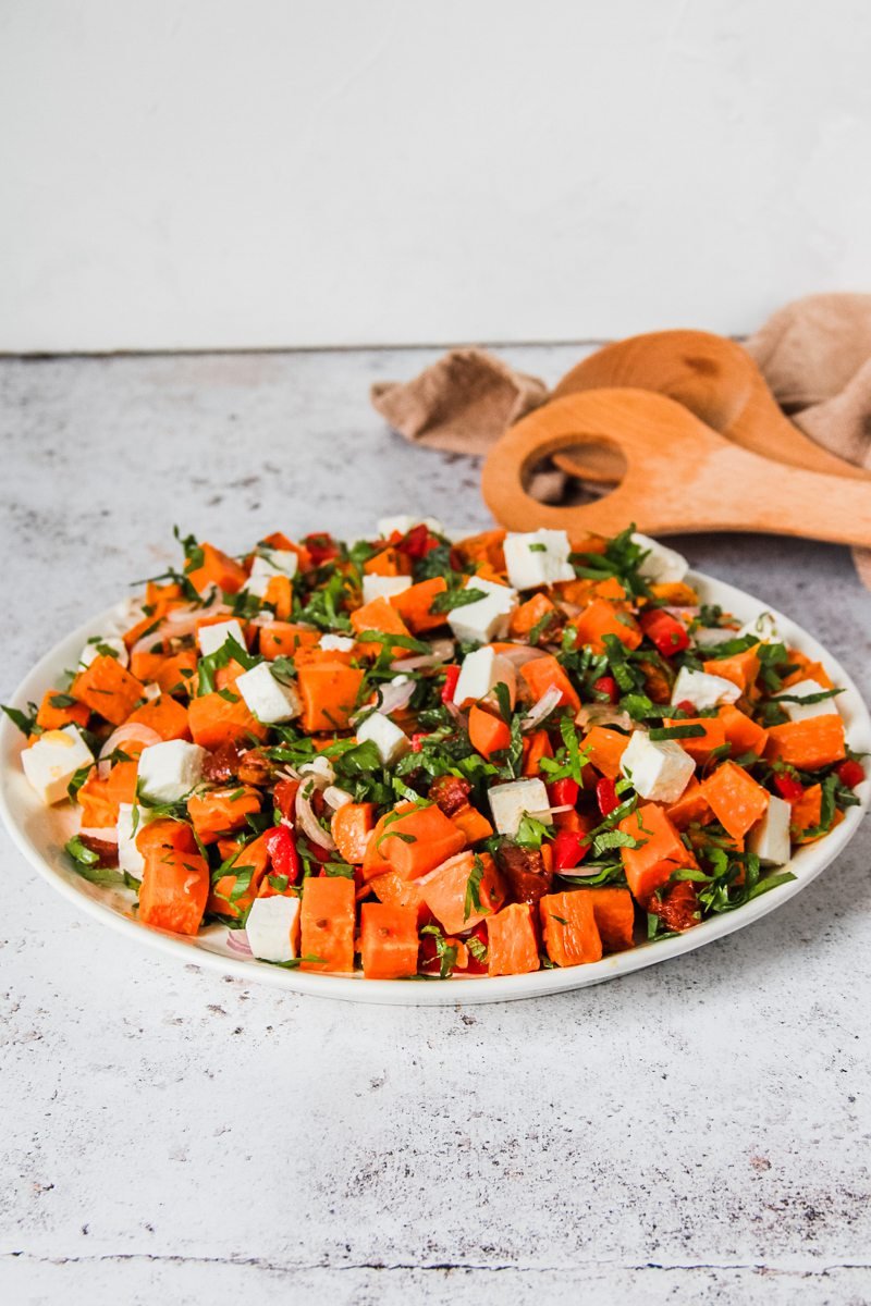 A sweet potato salad sits on a round white ceramic plate on a gray surface with salad servers on the side.