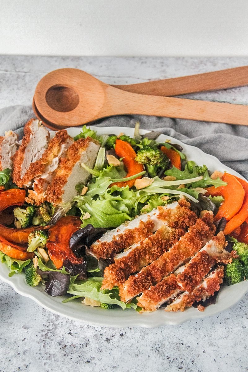 Crispy chicken slices sit on top of lettuce and roasted butternut squash served up on a white plate on a gray surface.