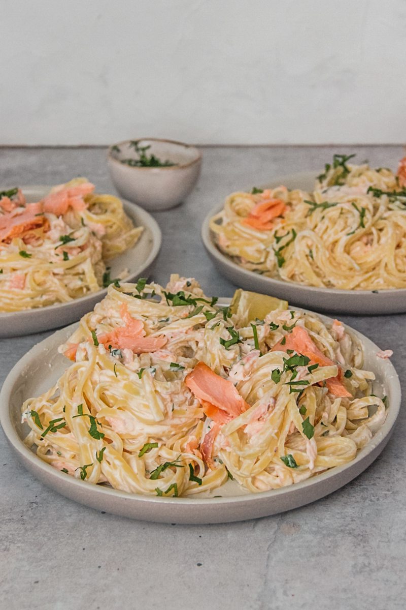 A side shot of rimmed gray ceramic plates with creamy smoked salmon pasta topped with finely chopped parsley and lemon wedges sitting on a gray surface.