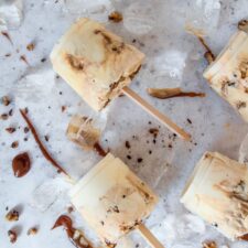 Salted Caramel Yogurt Popsicles sit on ice cubes on caramel drizzles on a light grey surface