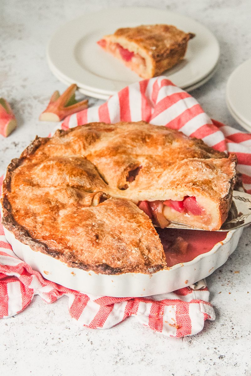 A slice of Irish rhubarb pie sits on a cake server as it is lifted from the remaining pie in a white ceramic baking dish on a striped cloth on a light gray surface.