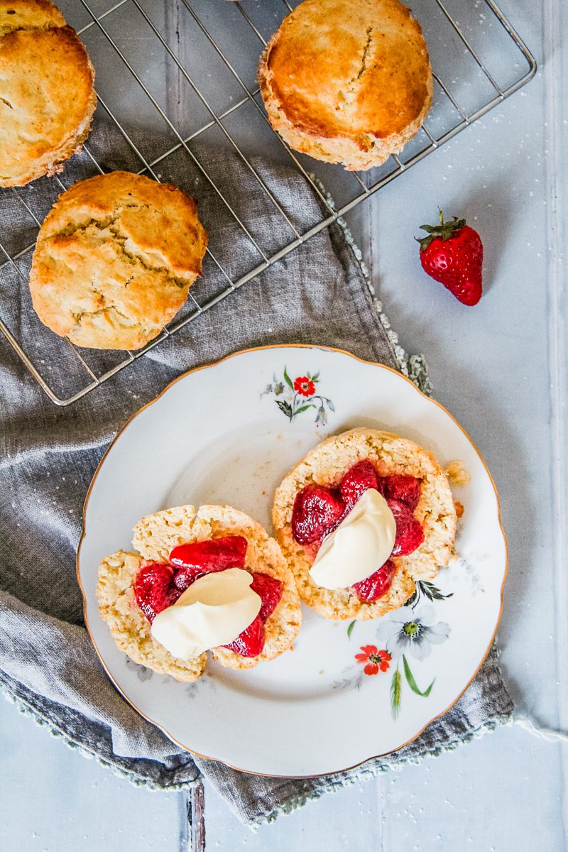 Halved scones topped with roasted strawberries and lightly whipped cream sit on a individual plate on a gray cloth and gray background with whole scones beside.