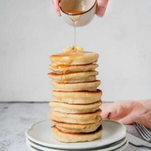 A stack of Scotch pancakes, also known as Griddle cakes, sits on a stack of plates being drizzled with honey on a light grey surface.