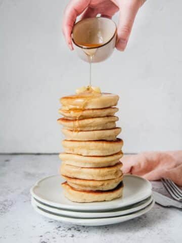 A stack of Scotch pancakes, also known as Griddle cakes, sits on a stack of plates being drizzled with honey on a light grey surface.