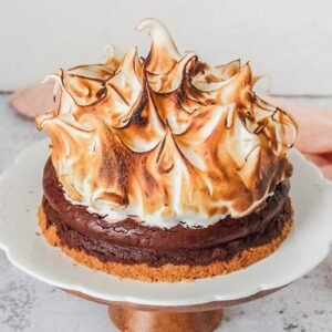 A smore pie sits on a white ceramic plate on a wooden cake stand on a light gray surface.