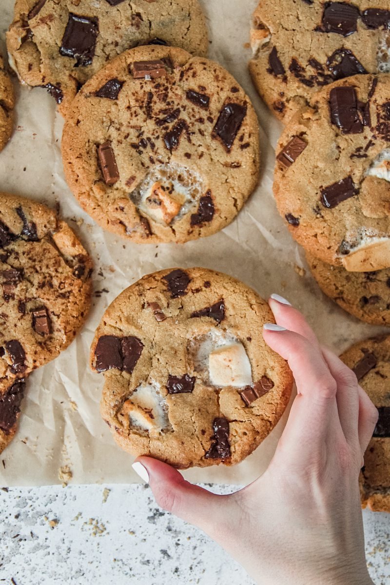 A Smore cookie is picked up from a light gray background with remaining cookies on parchment paper.