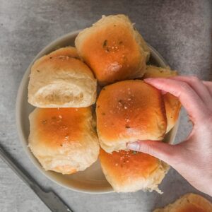 A dinner roll is picked up from a stack on a rimmed ceramic plate on a gray surface.