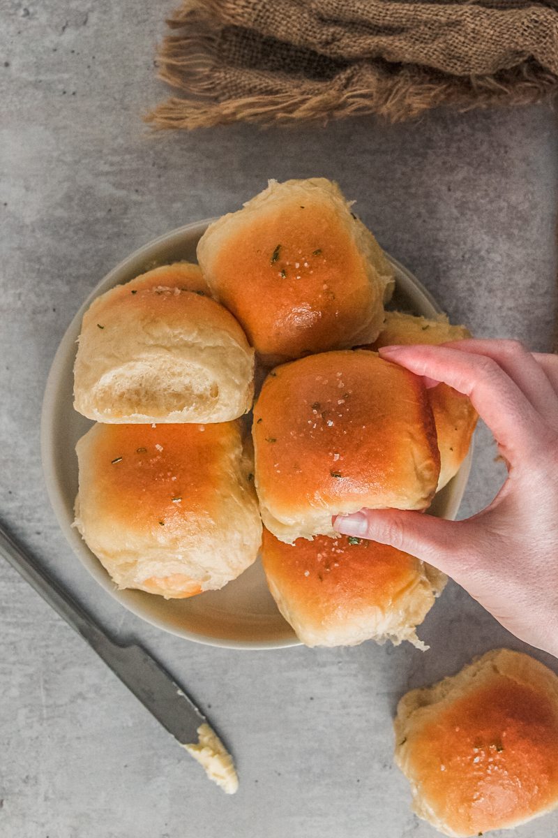 A dinner roll is picked up from a stack on a rimmed ceramic plate on a gray surface.