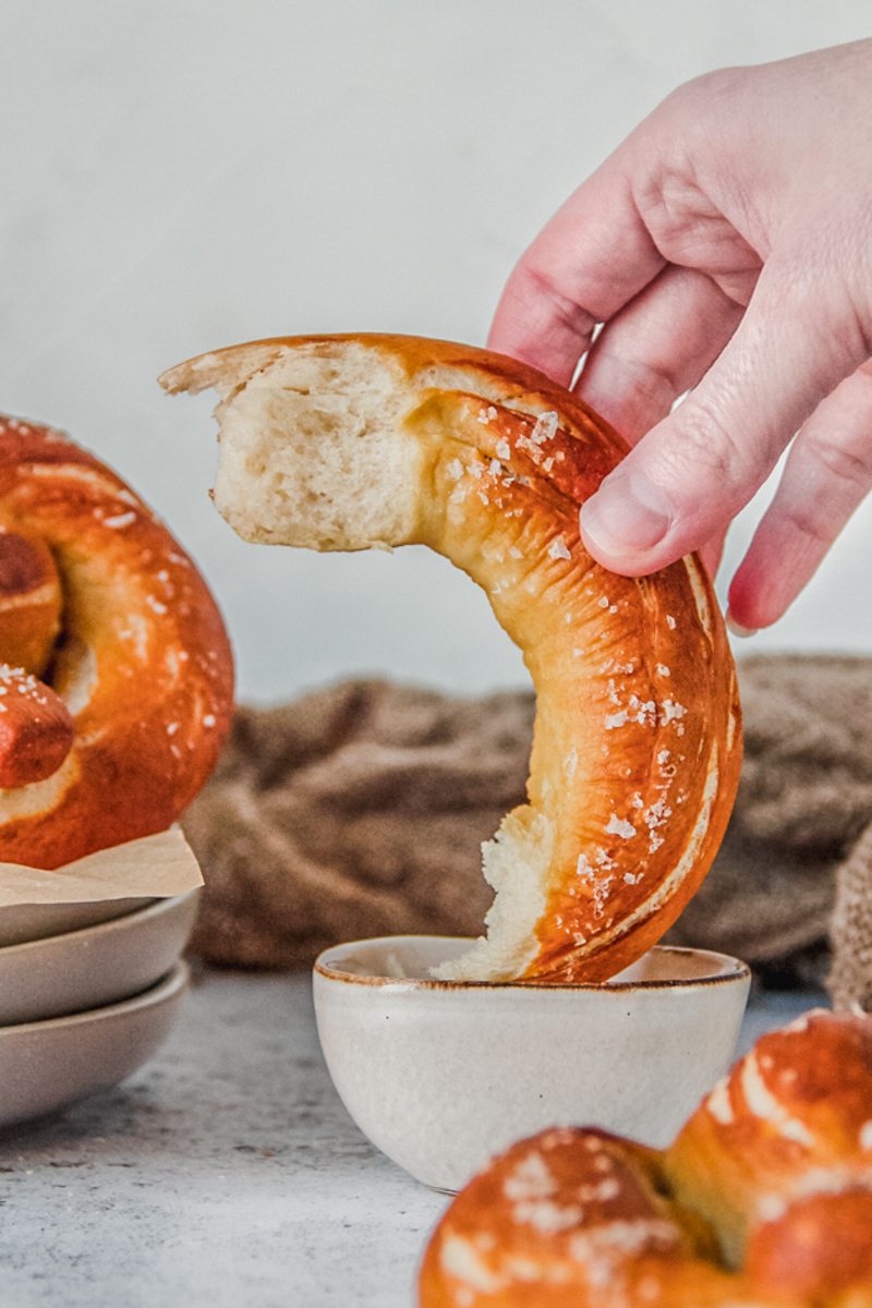 A piece of baked pretzel is dipped into mustard in a small bowl on a light gray surface.