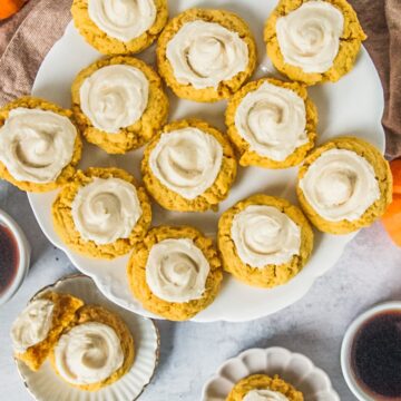 Pumpkin sugar cookies with maple buttercream frosting sit on a scalloped white ceramic plate above a light gray surface with individual plates and cookies beside.