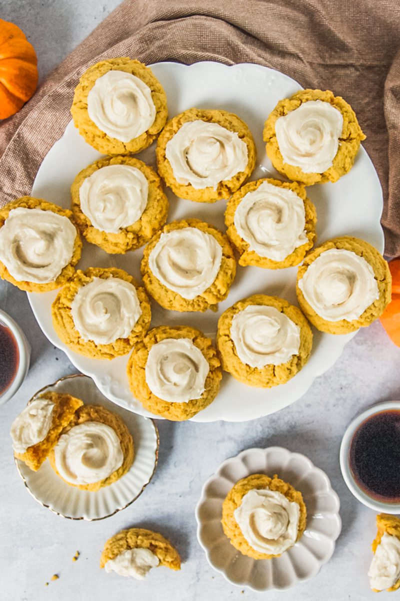 Pumpkin sugar cookies with maple buttercream frosting sit on a scalloped white ceramic plate above a light gray surface with individual plates and cookies beside.