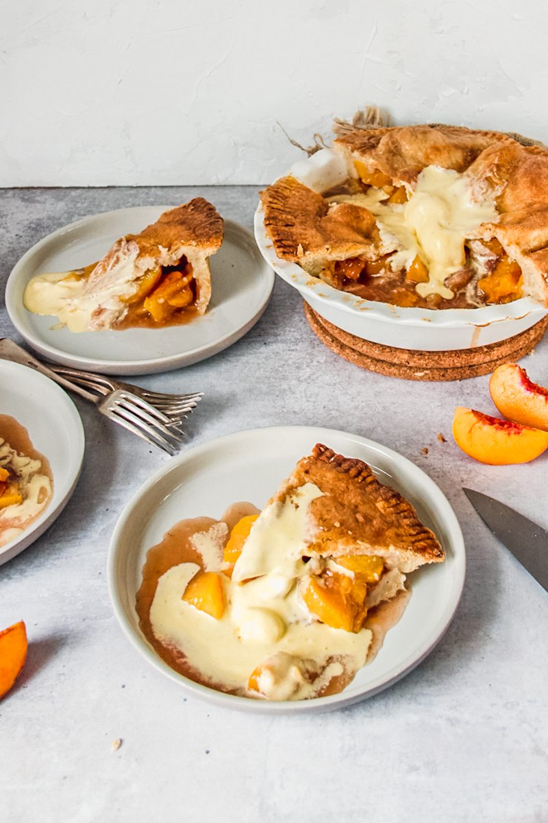 Individual servings of double crusted peach cobbler sit on plates with melting vanilla ice cream mixed with the peach juices beside serving forks and the remaining pie on a gray surface.