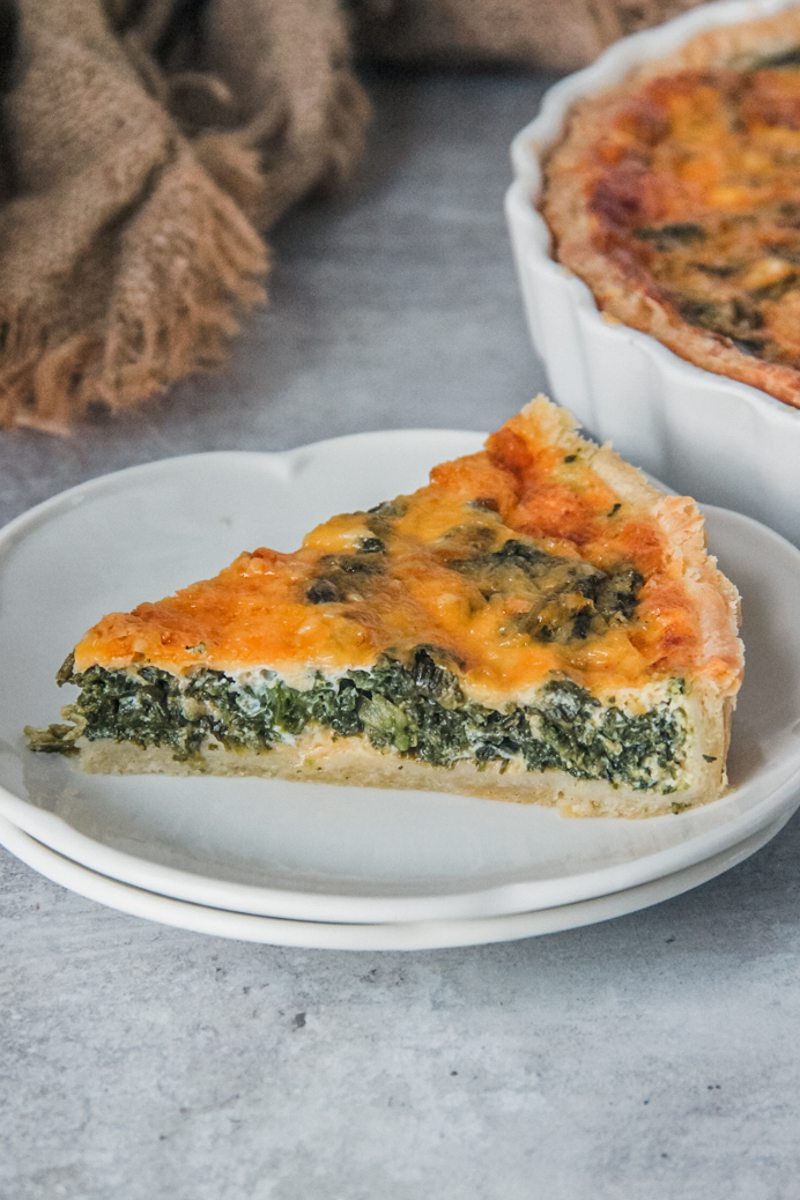 A slice of spinach and cheese quiche sits on a two white ceramic plates on a gray surface.