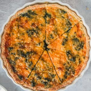 A spinach and cheese quiche sits in a white ceramic baking dish on a gray surface with slices cut through it.