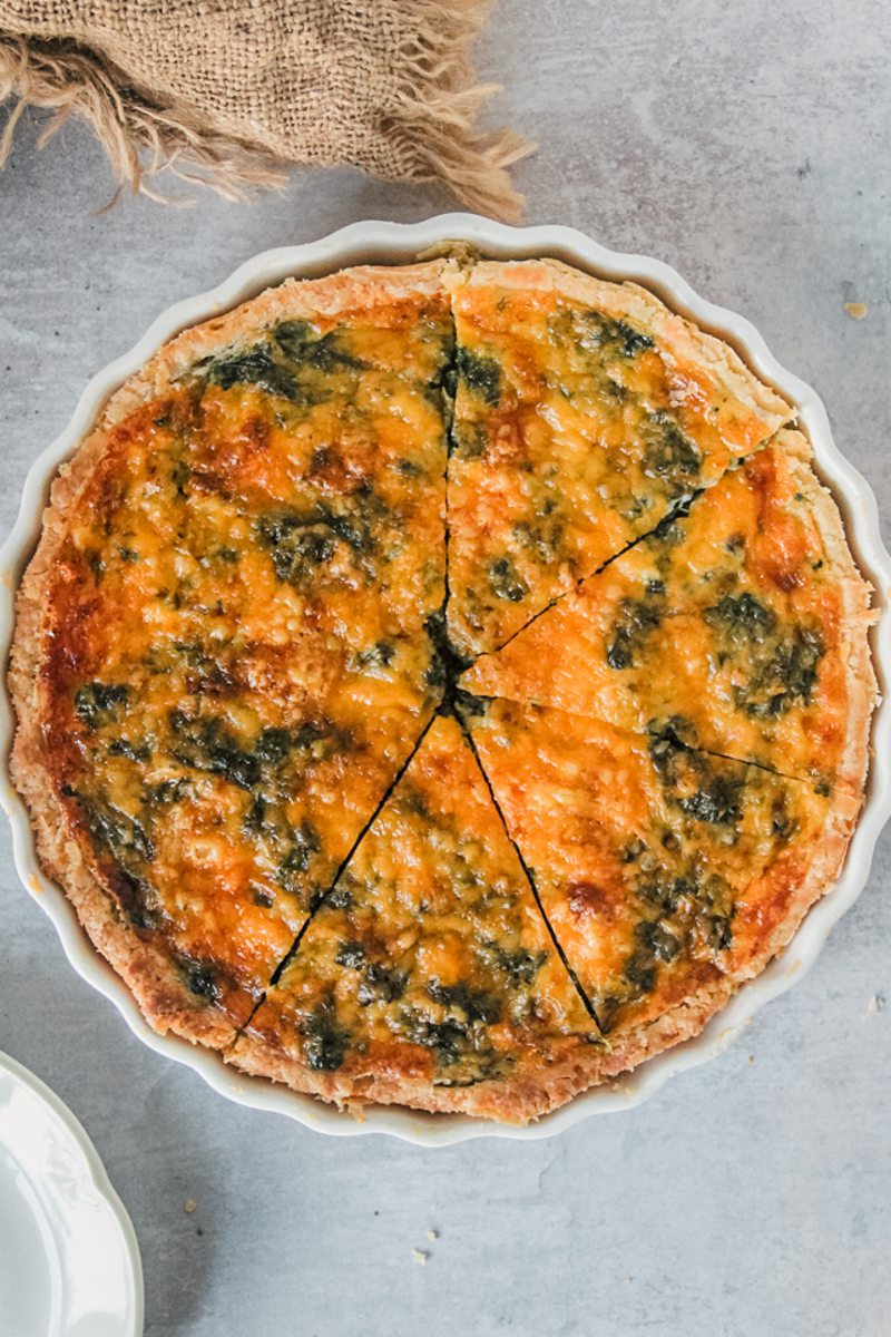 A spinach and cheese quiche sits in a white ceramic baking dish on a gray surface with slices cut through it.