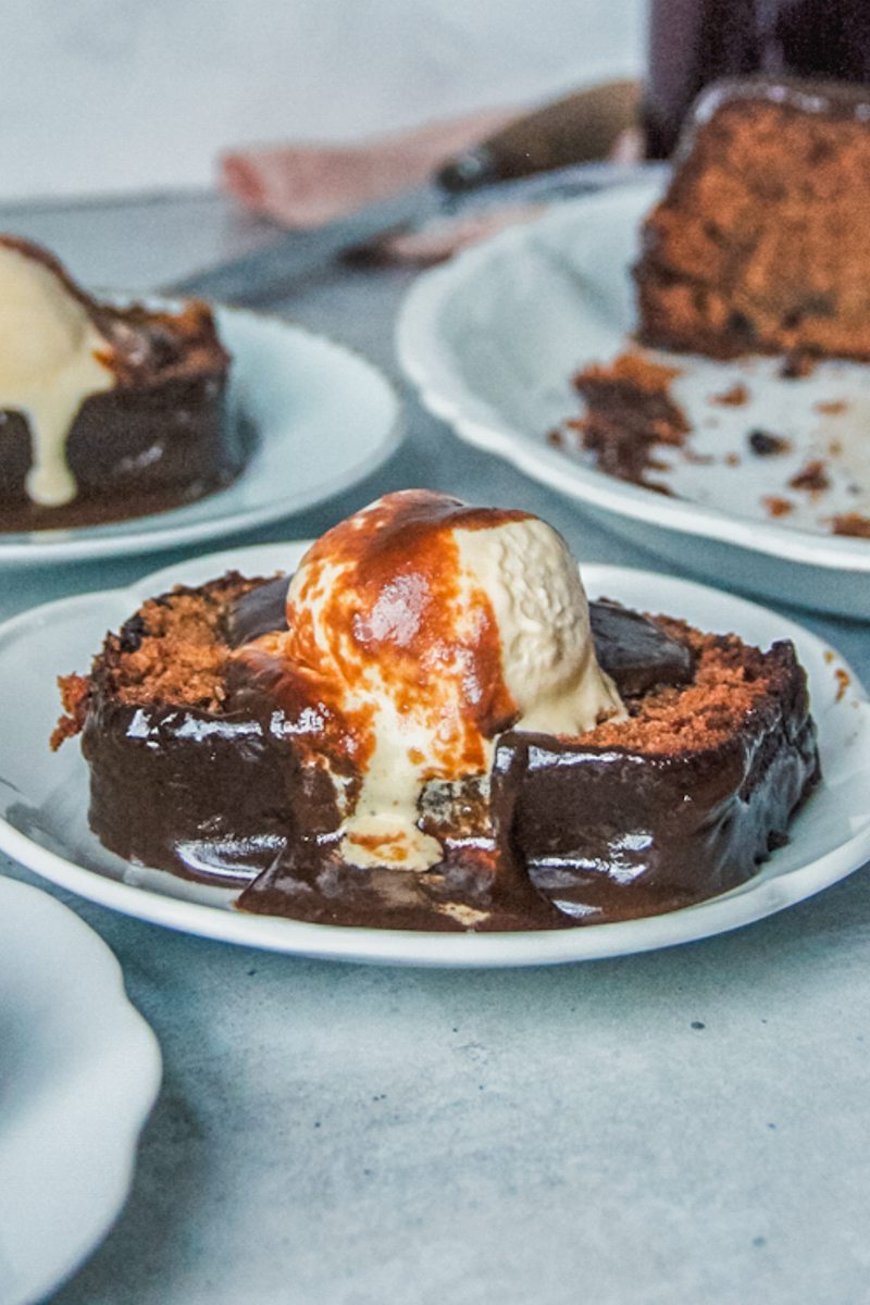 A slice of sticky toffee pudding cake sits on individual plates with a scoop of melting vanilla ice cream and toffee sauce on top on a gray surface.