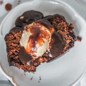 An overhead image of a slices of sticky toffee pudding sitting on individual plates with a scoop of melting vanilla ice cream and toffee sauce on top on a gray surface.