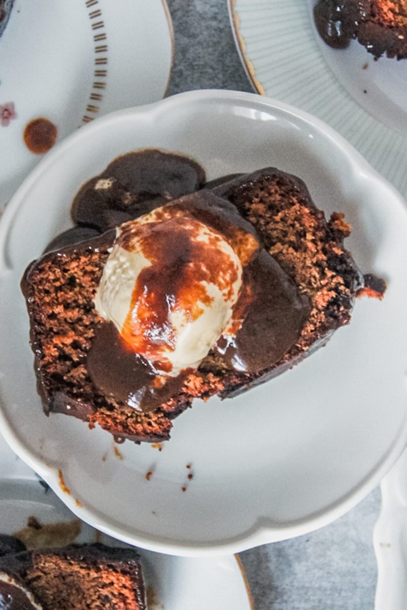 An overhead image of a slices of sticky toffee pudding sitting on individual plates with a scoop of melting vanilla ice cream and toffee sauce on top on a gray surface.