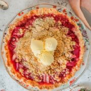 A Strawberry Rhubarb pie sits on a floral ceramic plate on a light gray surface with a few scoops of vanilla ice cream in the center with a pastel cloth beside.