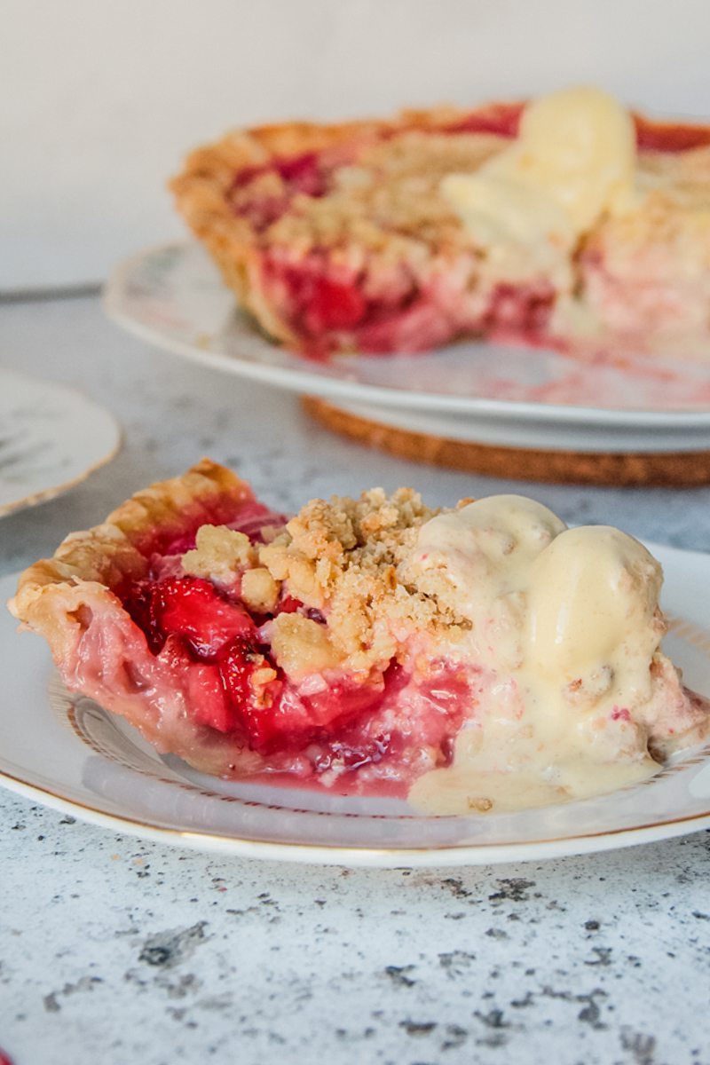 A slice of strawberry rhubarb pie sits on an individual plate with a scoop of vanilla ice cream on a light gray surface.