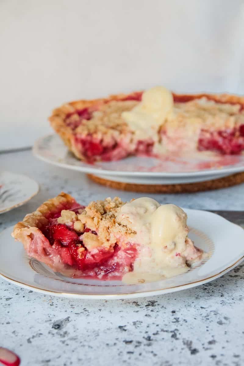 A slice of strawberry rhubarb crumble pie sits served up on a plate with a scoop of vanilla ice cream melting at the front of the slice, on a light grey background.