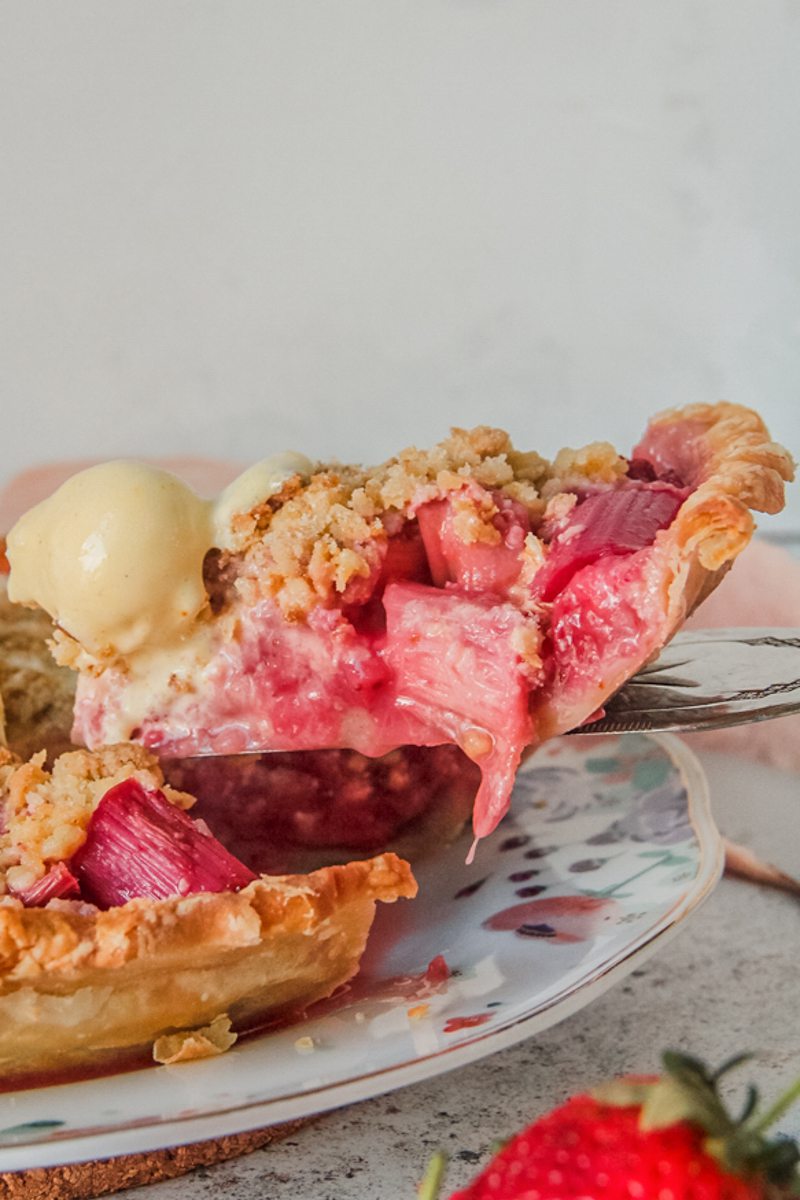 A slice of strawberry rhubarb pie sits on a server with a scoop of melting ice cream on top beside the remaining pie on a light gray surface.