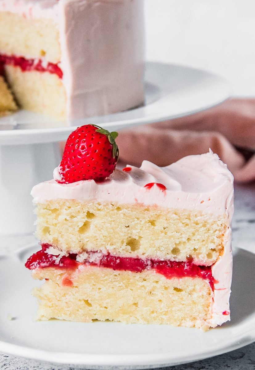 A close up of a slice of two layer vanilla cake with roasted strawberries in the center and coated in a strawberry buttercream, served up on a plate with the cake in the background.
