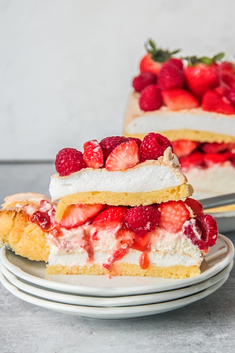 A slice of Swedish meringue cake with berries and cream sits on a stack of white ceramic plates on a gray surface.