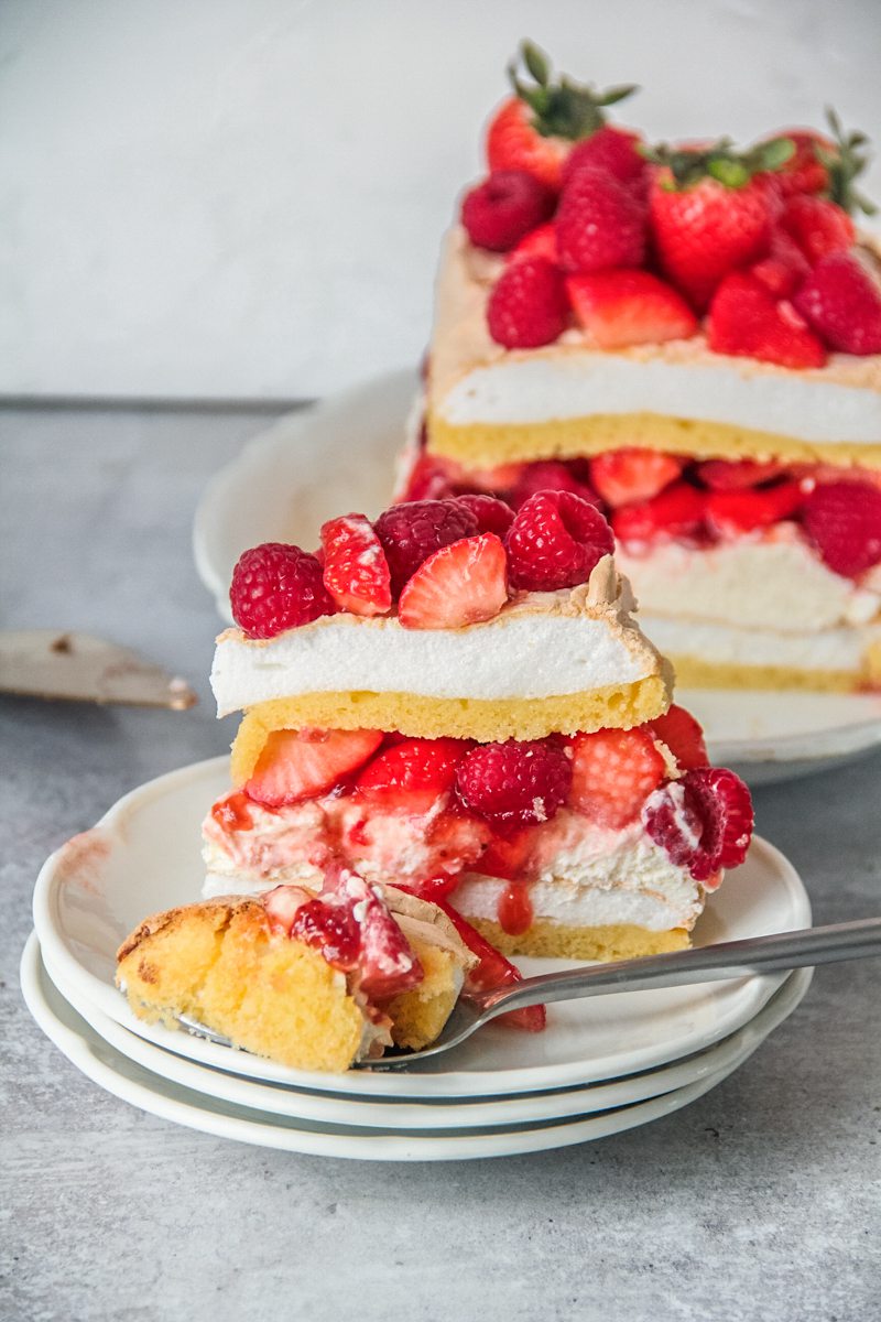 A slice of Swedish meringue cake with a spoonful resting on the edge of the plate sits on a stack of white ceramic plates on a gray surface.