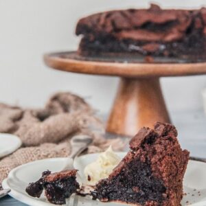 A slice of Fudgy Swedish Chocolate cake is served up on an individual white ceramic plate with it's gooey interior sitting on a fork beside a dollop of whipped cream on a light blue surface.