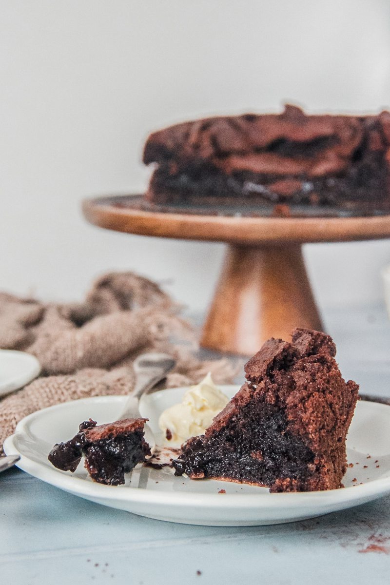 A slice of Fudgy Swedish Chocolate cake is served up on an individual white ceramic plate with it's gooey interior sitting on a fork beside a dollop of whipped cream on a light blue surface.