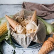 Scoops of no churn sweet corn ice cream sit inside waffle cones that sit rested on the round tin of ice cream surrounded by fresh corn and waffle cones on a light grey background.