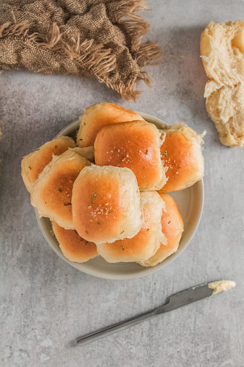 Dinner rolls sits stacked on a rimmed ceramic plate on a gray surface with a butter knife and a little butter on the side and a further dinner roll halved on the side.