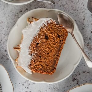 A slice of sweet potato loaf cake with a marshmallow topping sits on an individual serving plate on a light gray surface with a spoon beside.