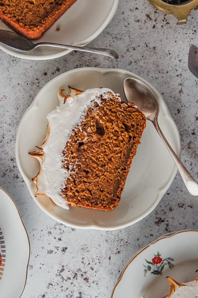 A slice of sweet potato loaf cake with a marshmallow topping sits on an individual serving plate on a light gray surface with a spoon beside.