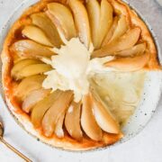 Pear tarte tatin topped up scoops of melting vanilla ice cream sits served up on a white plate on a gray surface.