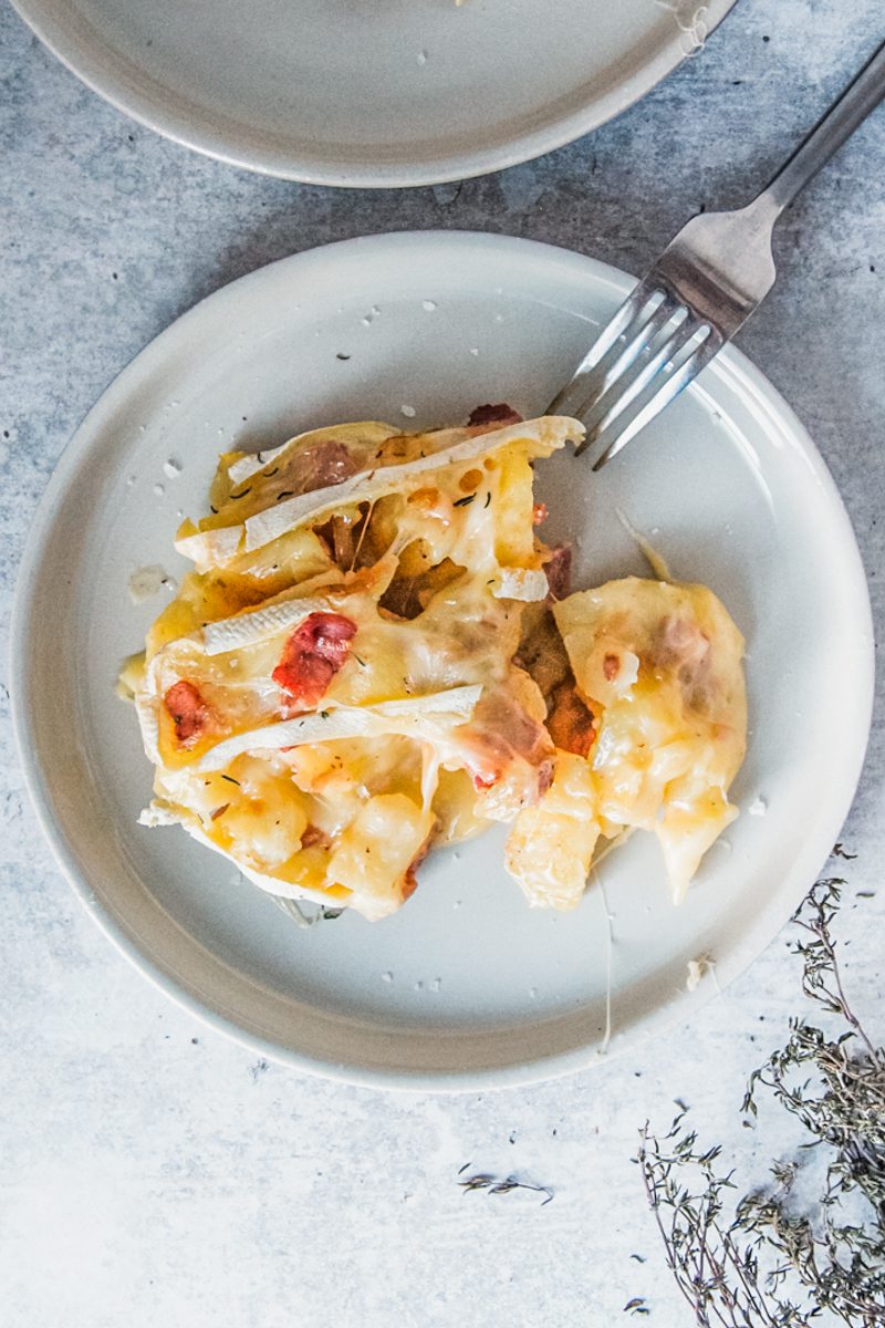 An individual serving of Tartiflette sits on a rimmed ceramic plate on a light gray surface with a fork resting on the edge of the plate.